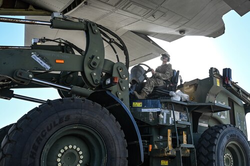 A giant, heavy-duty forklift operated by a man in uniform moves cargo on a flightline.