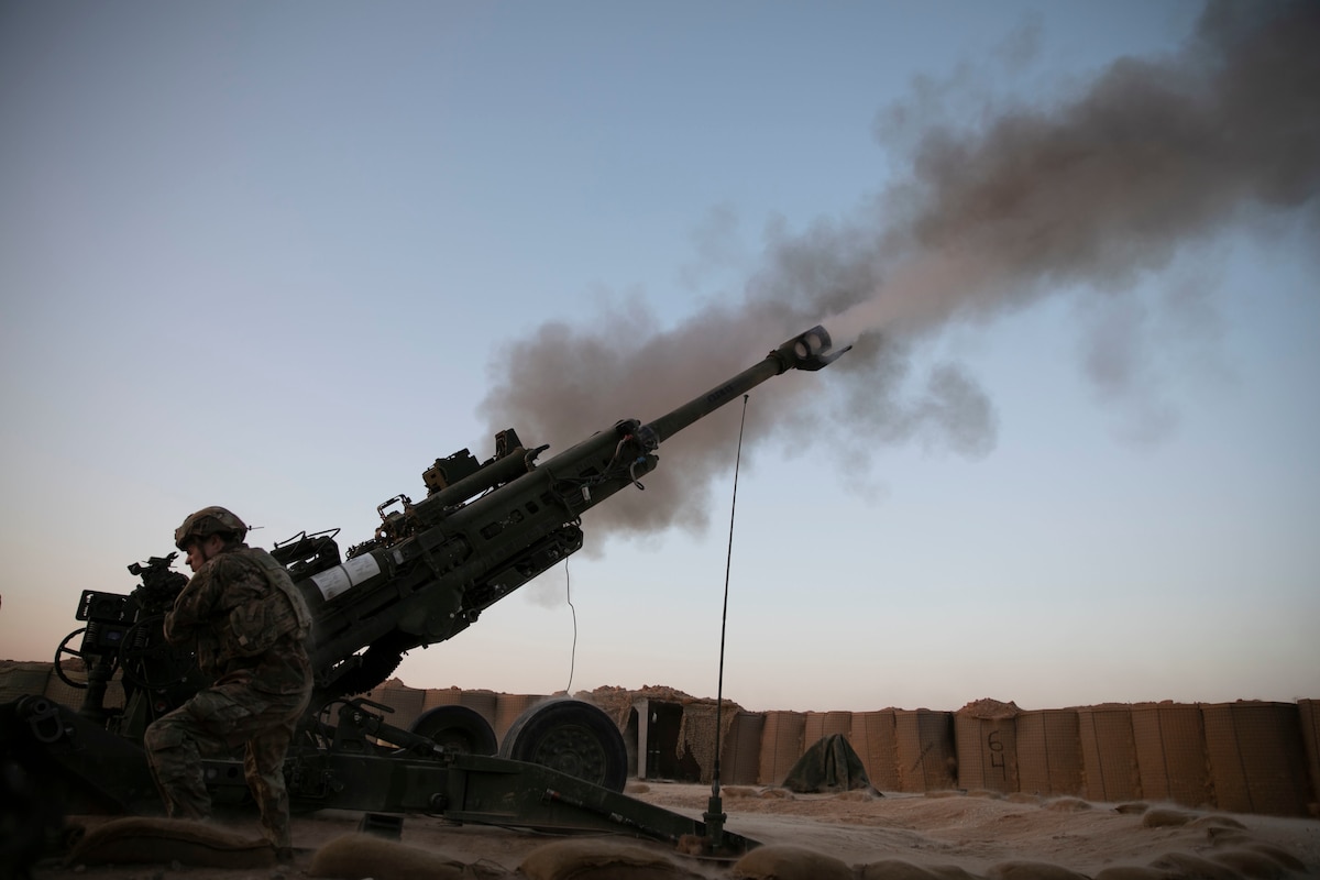 A soldier stands near the base of a large artillery weapon as it is being fired with minimal light into the sky.
