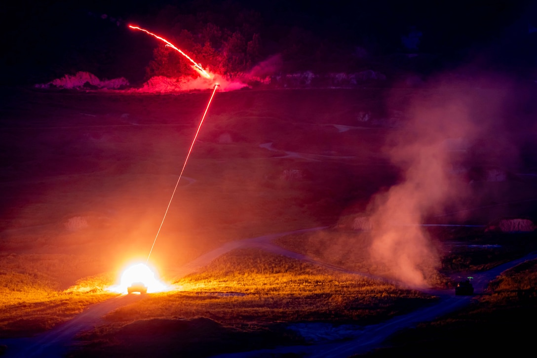 A tank fires a missile at night whose tail lights up the sky and the surrounding area.