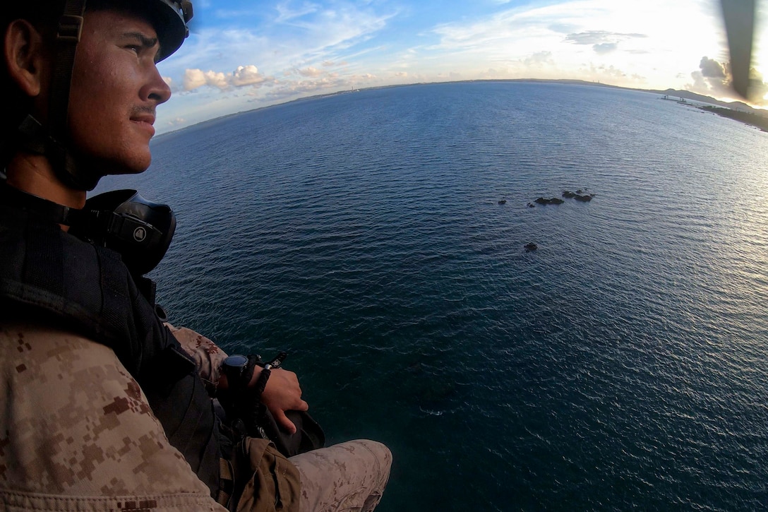 A Marine looks out at a sunny horizon from a helicopter flying over water.