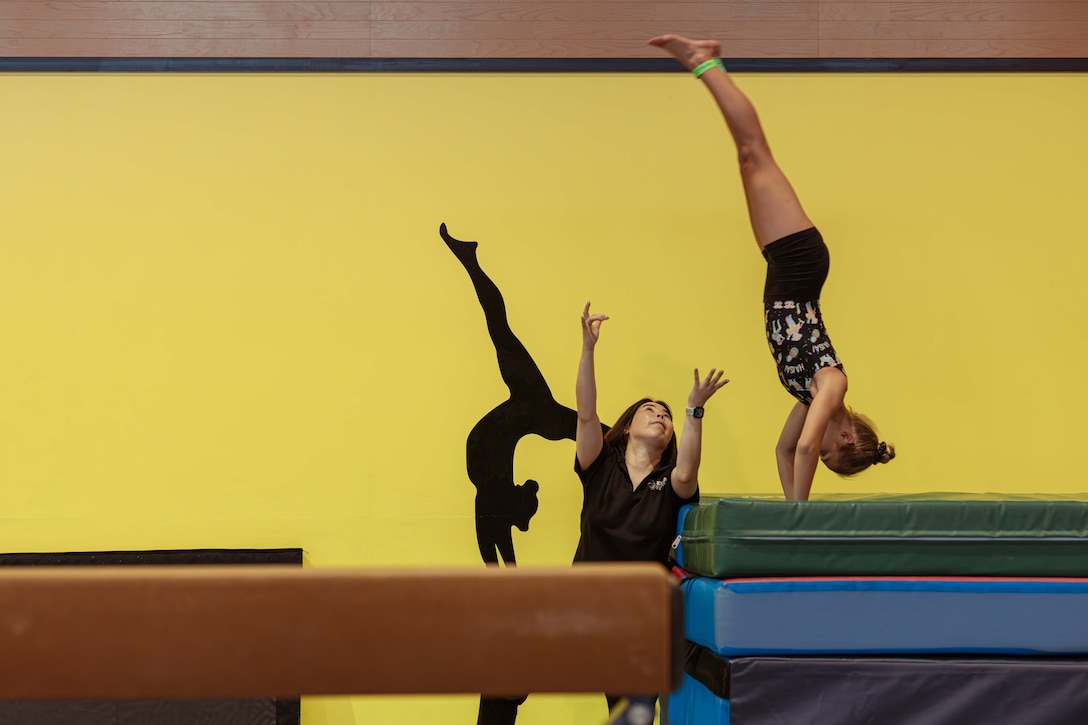 A coach spots a gymnast doing a handstand while performing a vault in a fitness center. The shadow of another gymnast is seen on a yellow wall in the background.