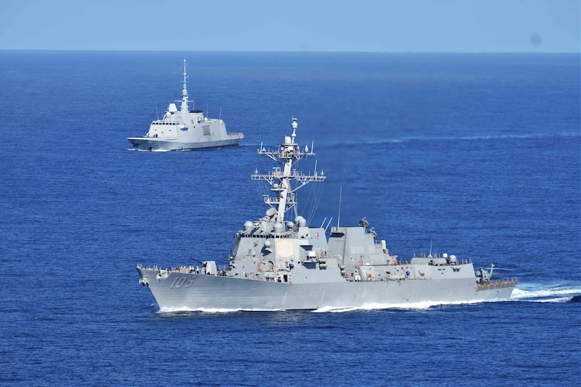 Two military ships steam close together in the ocean.
