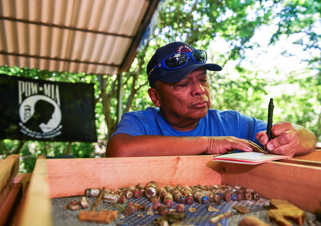 A man writes in a notebook. Behind him is a POW/MIA flag. In front of him is a wooden tray that contains bottles and other debris.