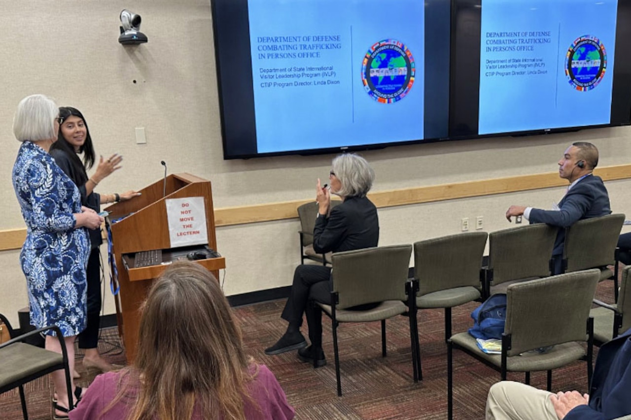 Two people at a lectern speak to a classroom audience with two large monitors on the wall.