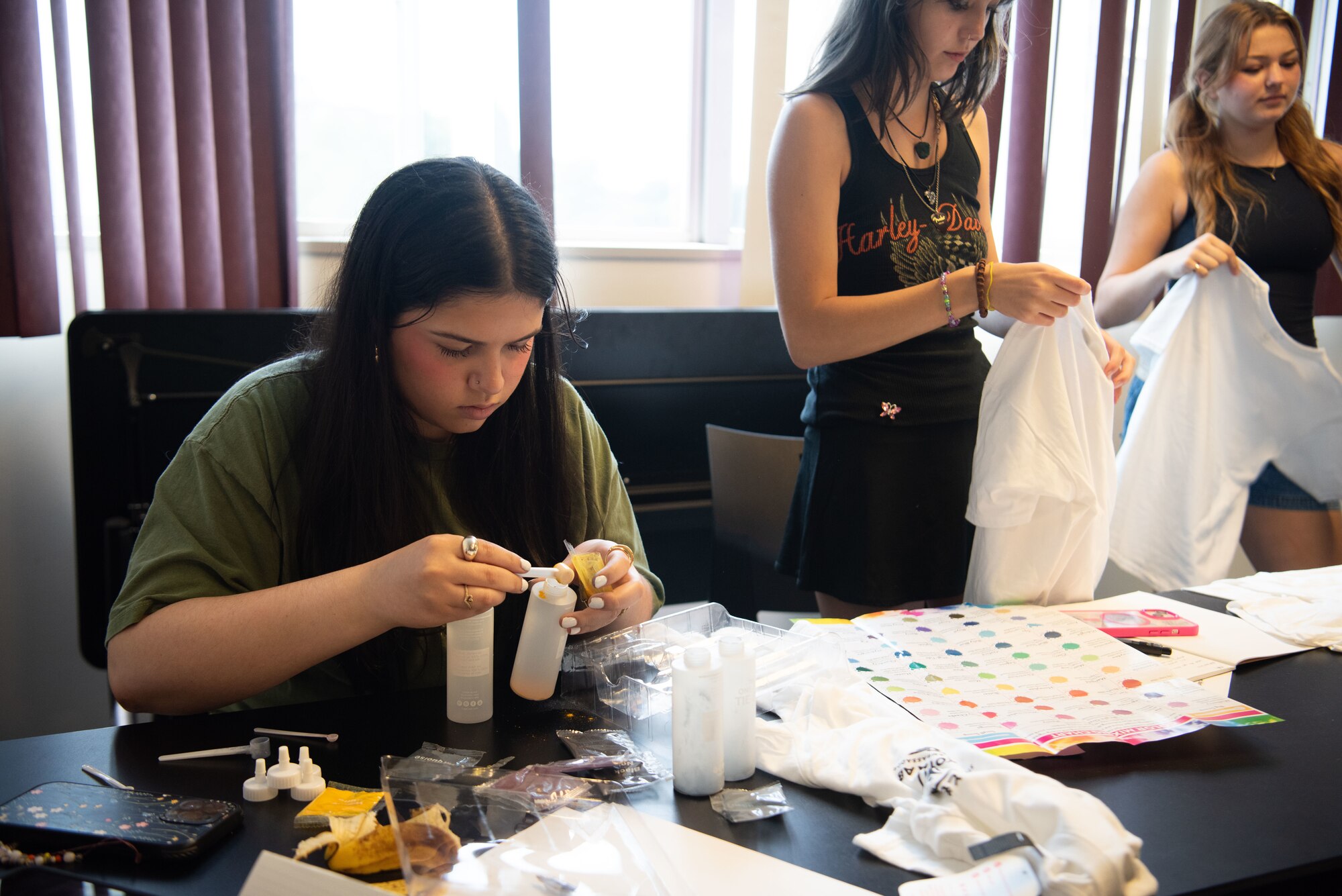 A student mixes die into a small container while two other students hold up white shirts in the background.
