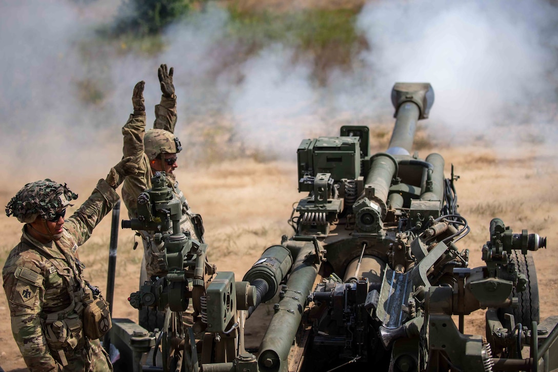 Two soldiers stand next to a howitzer, one with both arms raised, as smoke swirls through the air.