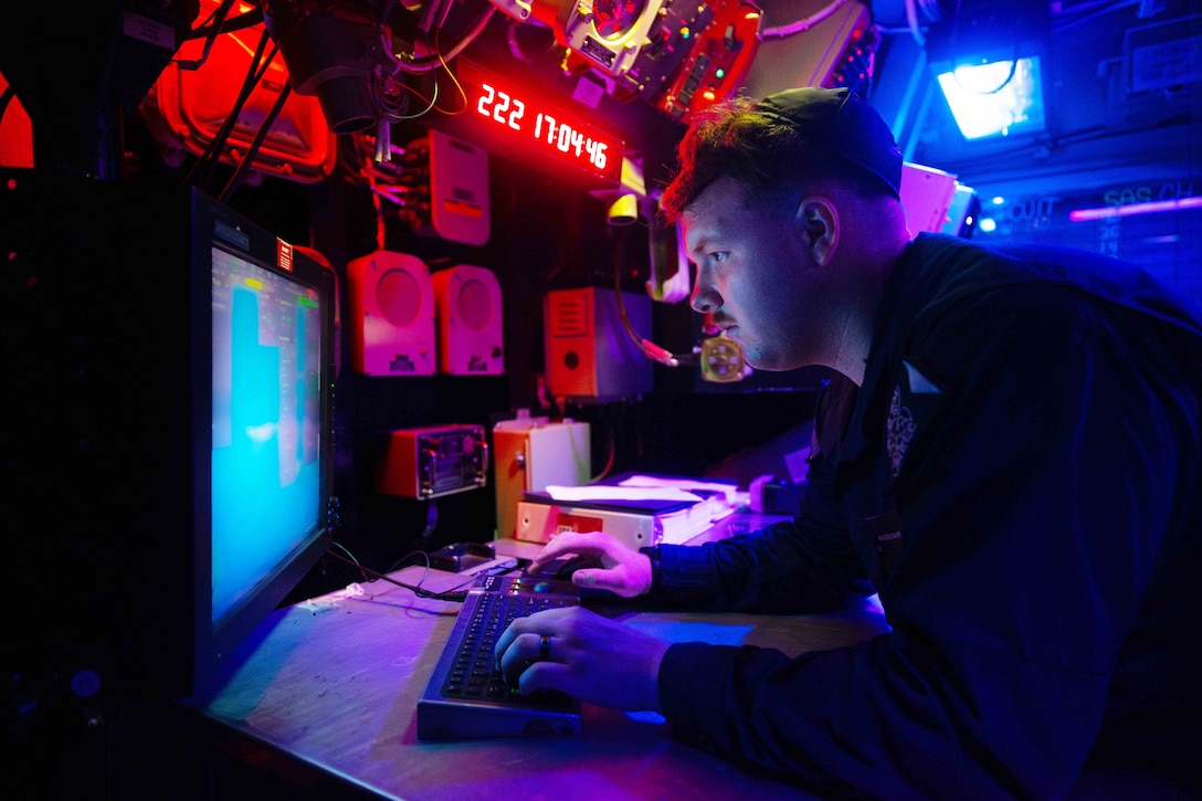 A sailor leans over a desk with hands on a keyboard while focusing on a monitor in a dark room with red and blue lighting.