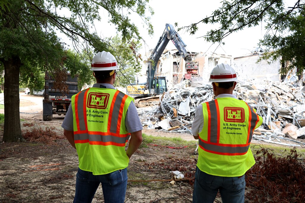 Two men in hard hats watch as a back hoe destroys an old facility.