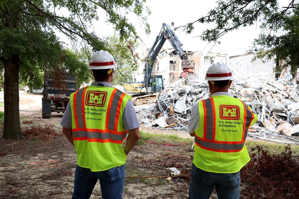 Two men in hard hats watch as a back hoe destroys an old facility.