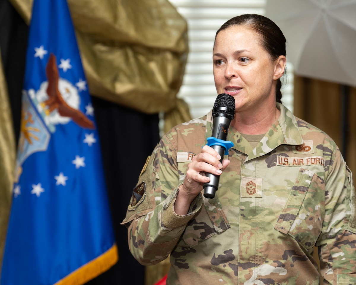 A woman in camouflage unform uses a handheld microphone, An Air Force flag stands on the left.