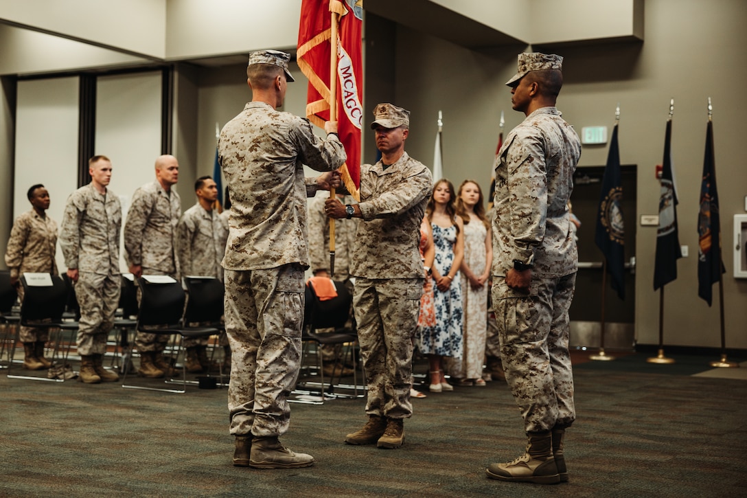 U.S. Marine Corps Lt. Col. Clinton Gebke, a San Antonio, Texas native, outgoing commanding officer of Headquarters Battalion, Marine Air-Ground Task Force Training Command, Marine Corps Air-Ground Combat Center, left, passes on the colors to Lt. Col. Nathan Rollins, a Huntsville, Texas native, incoming commanding officer of HQBN, center, during a HQBN change of command ceremony at MCAGCC, Twentynine Palms, California, July 23, 2024. The change of command ceremony represents the official passing of authority from the outgoing HQBN commander, Gebke, to the incoming commander, Rollins. (U.S. Marine Corps photo by Lance Cpl. Richard PerezGarcia)