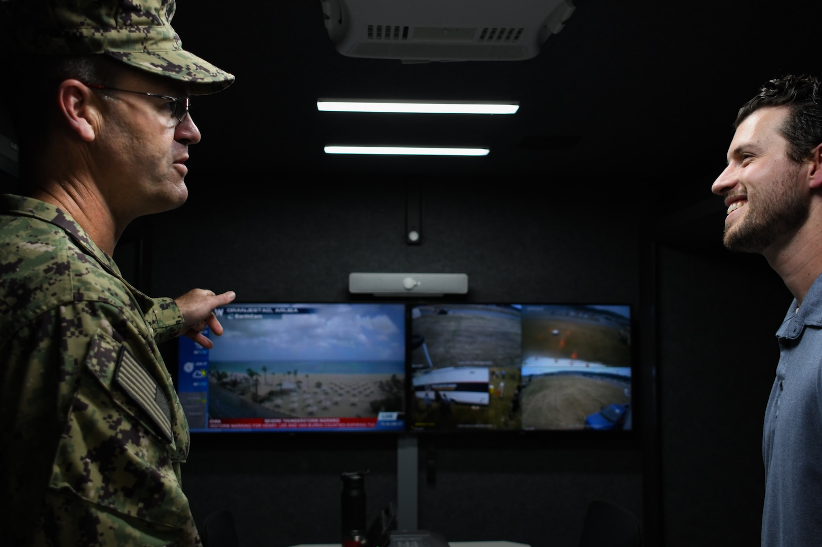 A Navy officer dressed in camouflage points at a tv screen and another man smiles