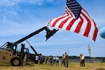 A group of people stand outside under an American flag under a blue sky on a grass field