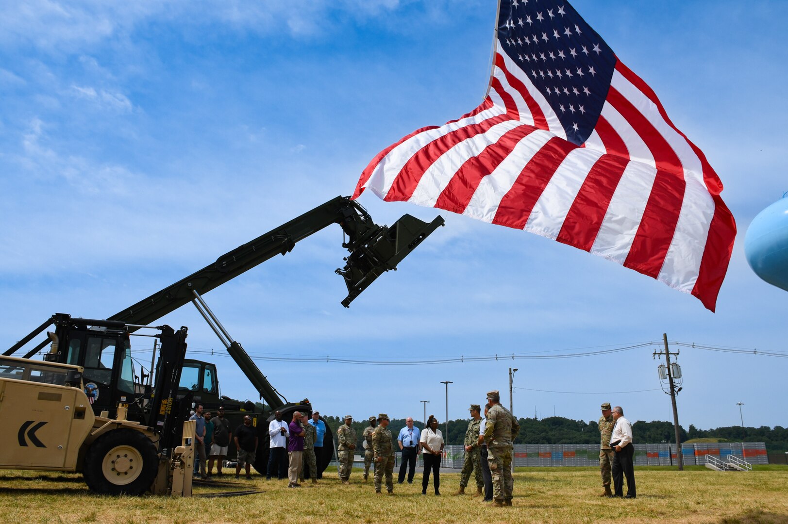 A group of people stand outside under an American flag under a blue sky on a grass field