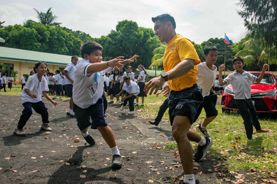 A group of excited schoolchildren chase a smiling sailor and play tag outside with the mountains, a red car and a school in the background.