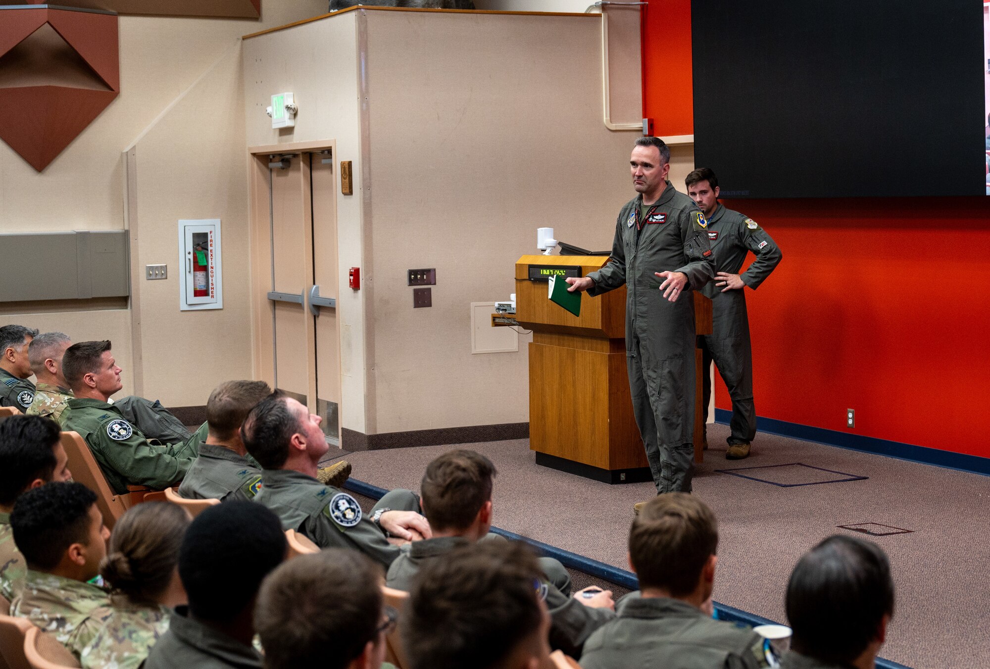 Two briefers stand on a stage to brief a crowd of seated people for the Red Flag-Alaska 24-3 exercise.