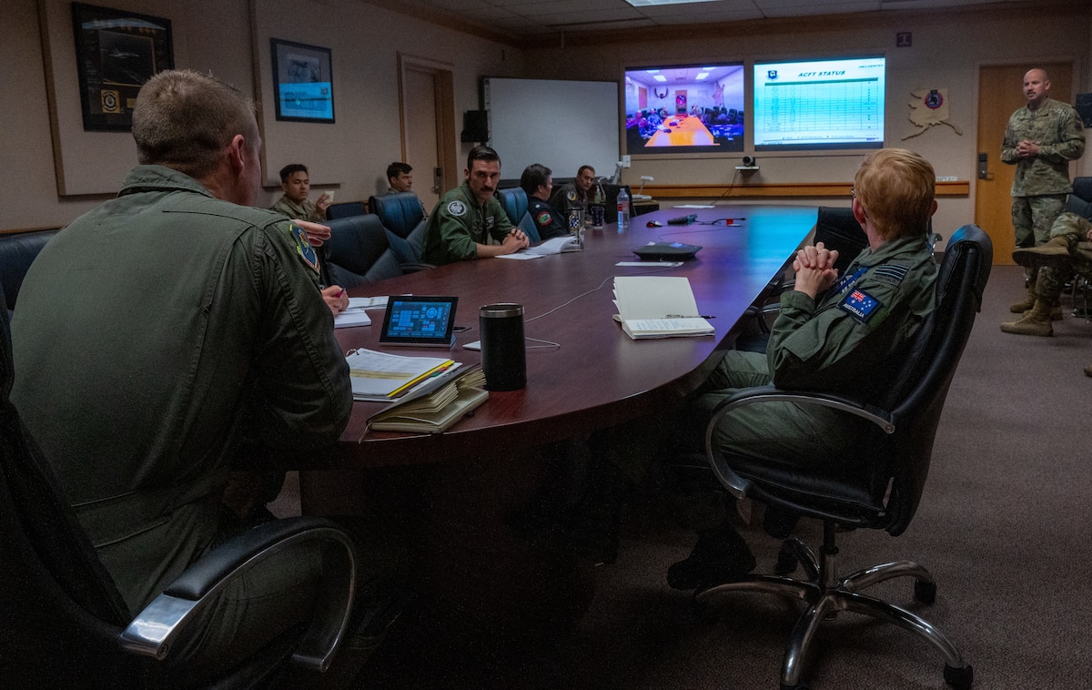 People sit around a wooden oval desk and listen to a presentation being shown on a projector in the front of the room.