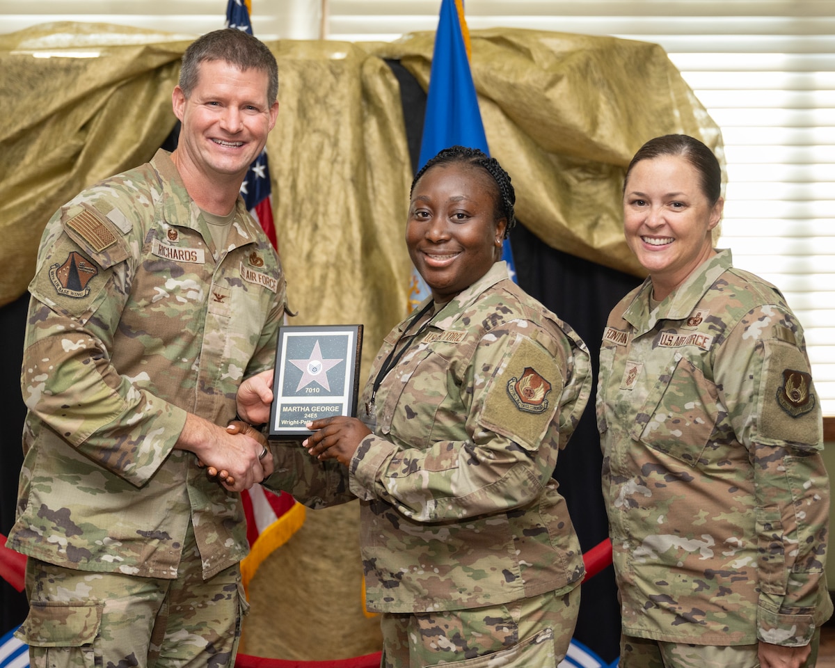 A man, on the left, shakes the hand of and presents a plaque to an African American woman. Another woman stand on the left. All of them are in camouflage uniforms