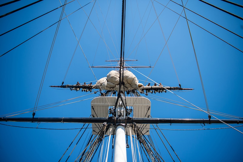 A group of sailors hang over the mast, as seen from below, under a blue sky.