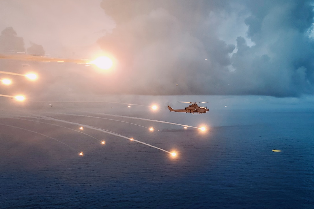 A military helicopter flies over open water against a cloudy sky as flares light up the sky.