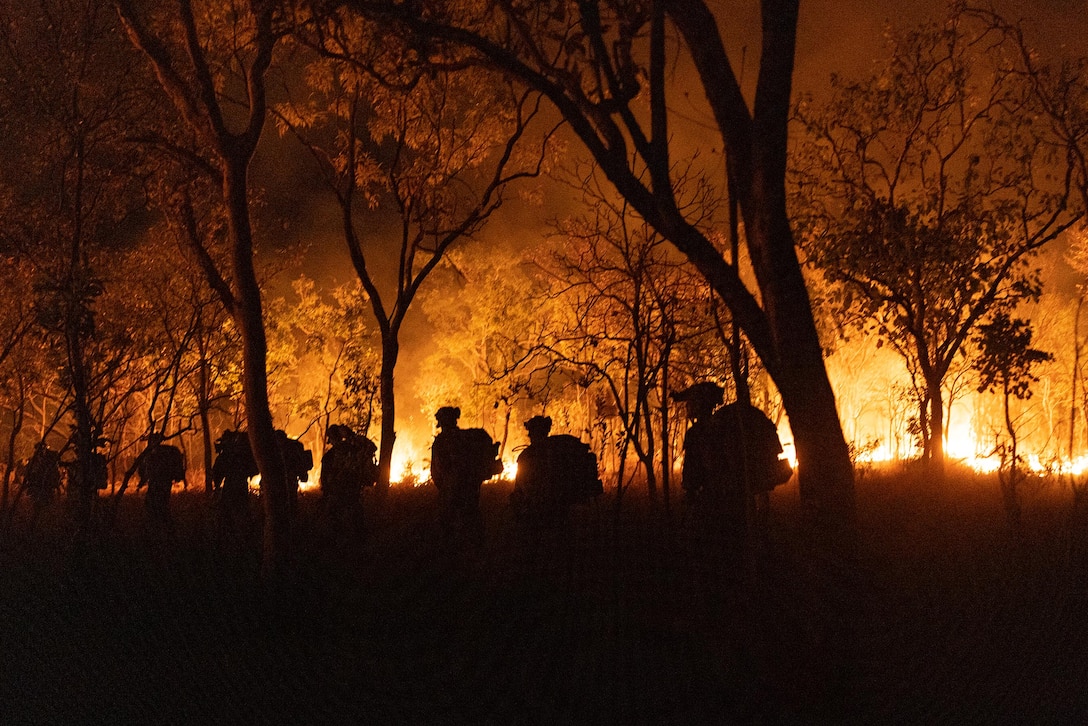 Silhouettes of Marines are seen patrolling a wooded area at night as fire burns in the background.