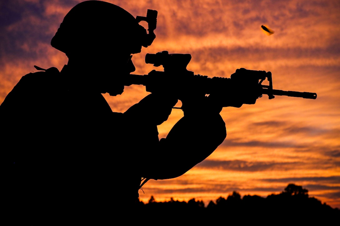 A bullet spirals in the air after a Marine fires a weapon under a sunlit sky as seen in silhouette.