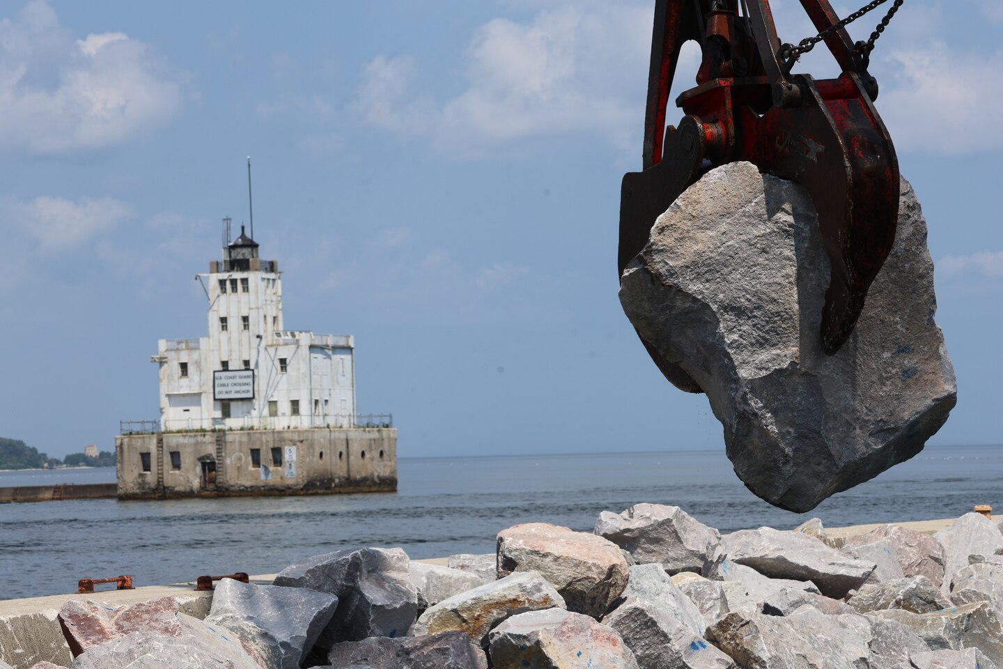 Crane is lifted high holding a huge boulder. The background shows an old lighthouse.