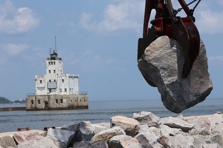 Crane is lifted high holding a huge boulder. The background shows an old lighthouse.