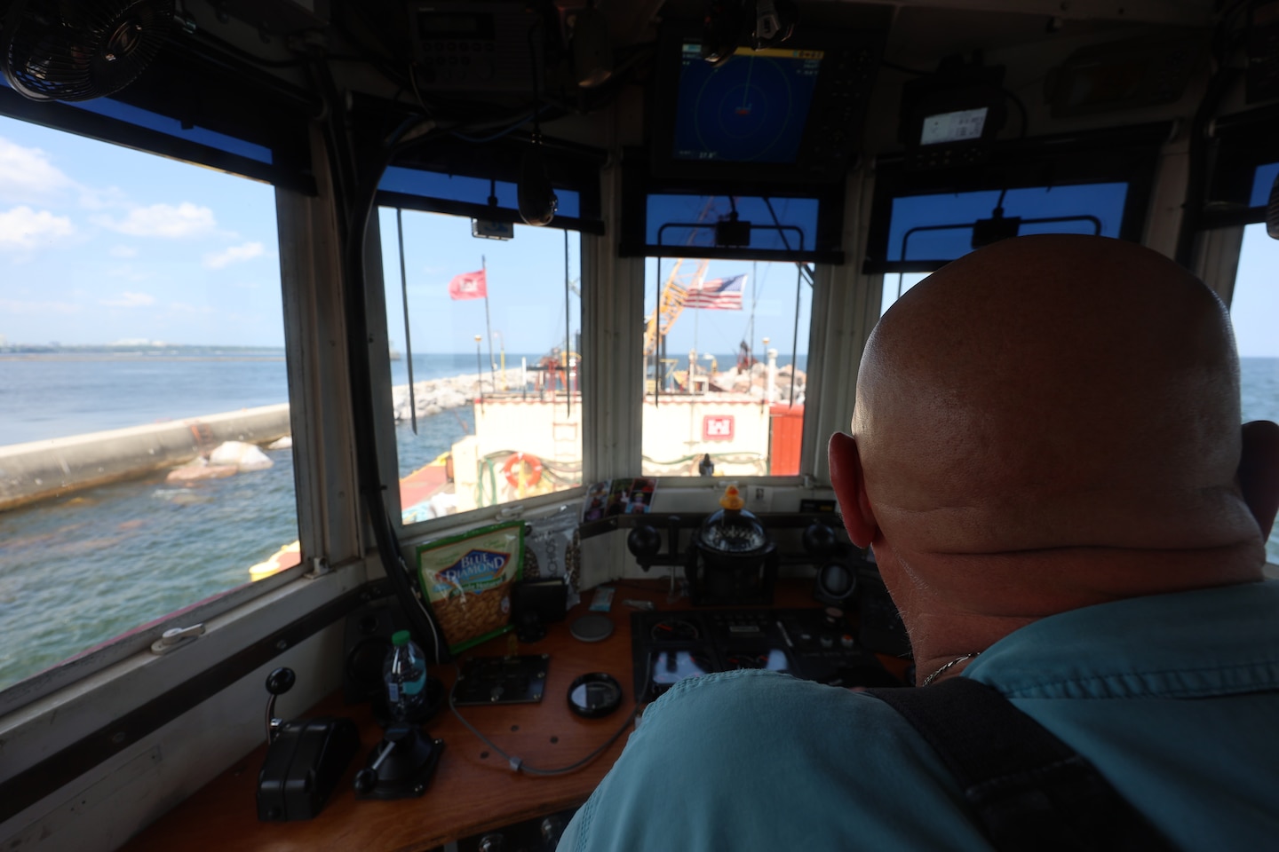 Man sits behind the wheel of a tugboat watching construction through the glass window.