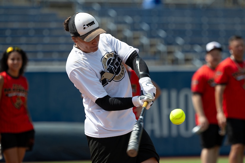 A woman wearing a softball uniform holds a bat and prepares to swing at a softball.