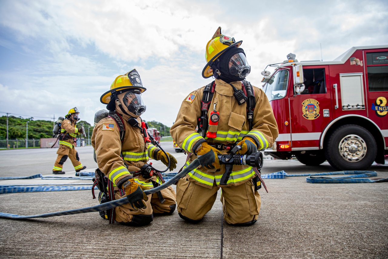 Firefighters in firefighting equipment kneel while holding a hose.