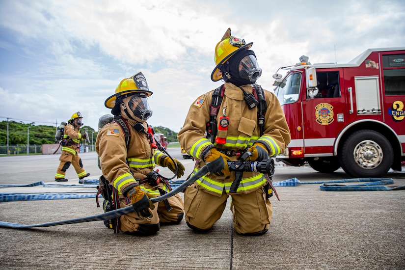 Firefighters in firefighting equipment kneel while holding a hose.