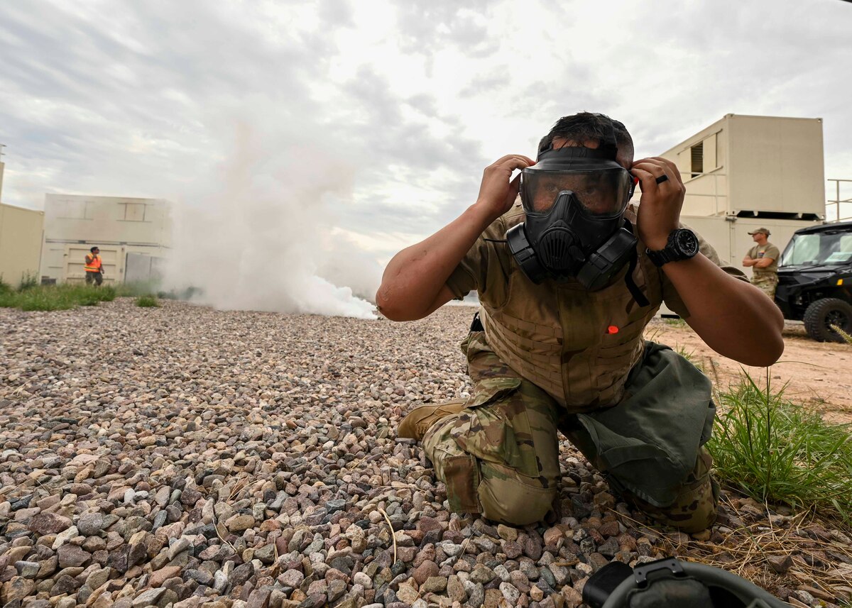 A man in a military uniform puts on a gas mask as white smoke approaches from behind.