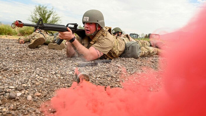 A man in a military uniform with a rifle lays next to red smoke.