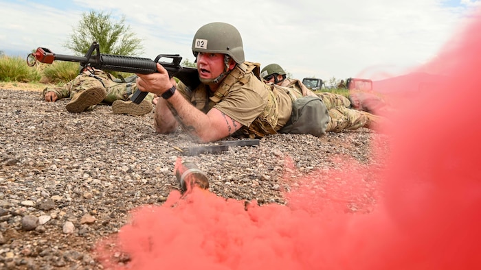 A man in a military uniform with a rifle lays next to red smoke.