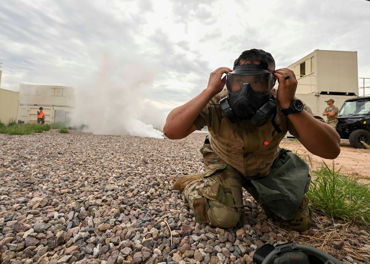 A man in a military uniform puts on a gas mask as white smoke approaches from behind.