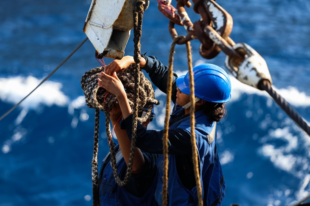 Two sailors wearing helmets work together with cables and a large amount of rope above the ocean in the background.