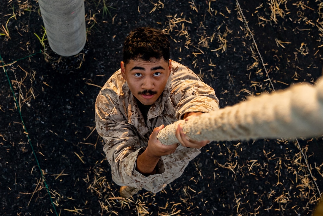 A Marine looks up at the camera while climbing a thick rope.