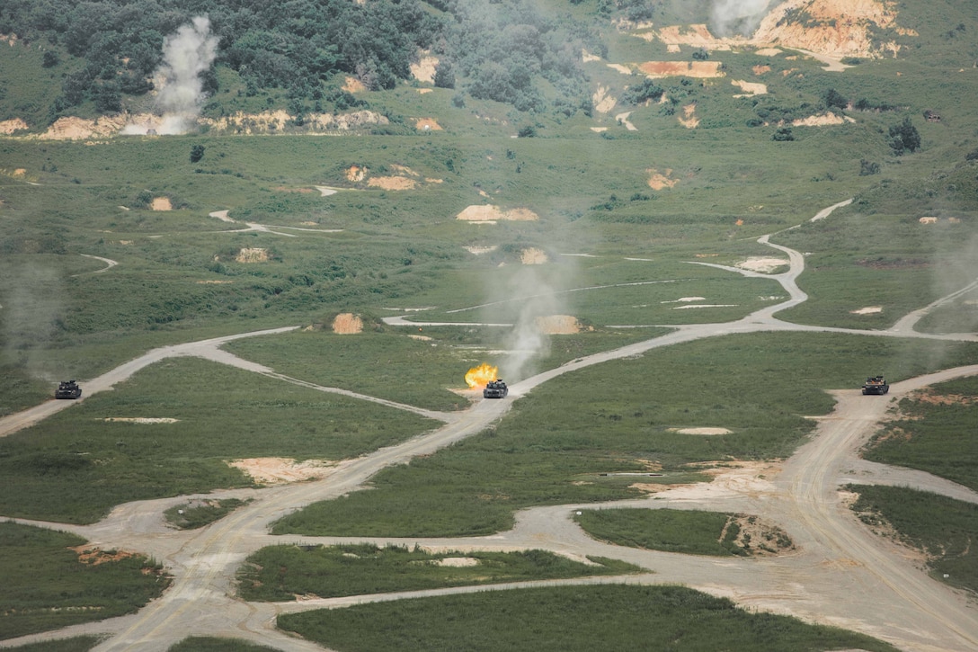 Three tanks travel along parallel dirt roads in a large open area. One of the tanks fires its weapon causing an explosion.