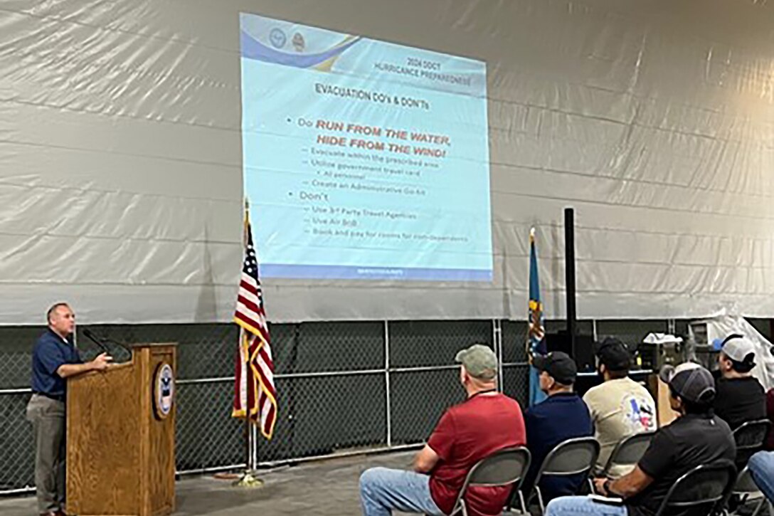 A man speaks to a group of seated people from behind a podium. There is a screen in front of the group that has hurricane safety information on it.