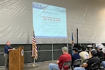 A man speaks to a group of seated people from behind a podium. There is a screen in front of the group that has hurricane safety information on it.