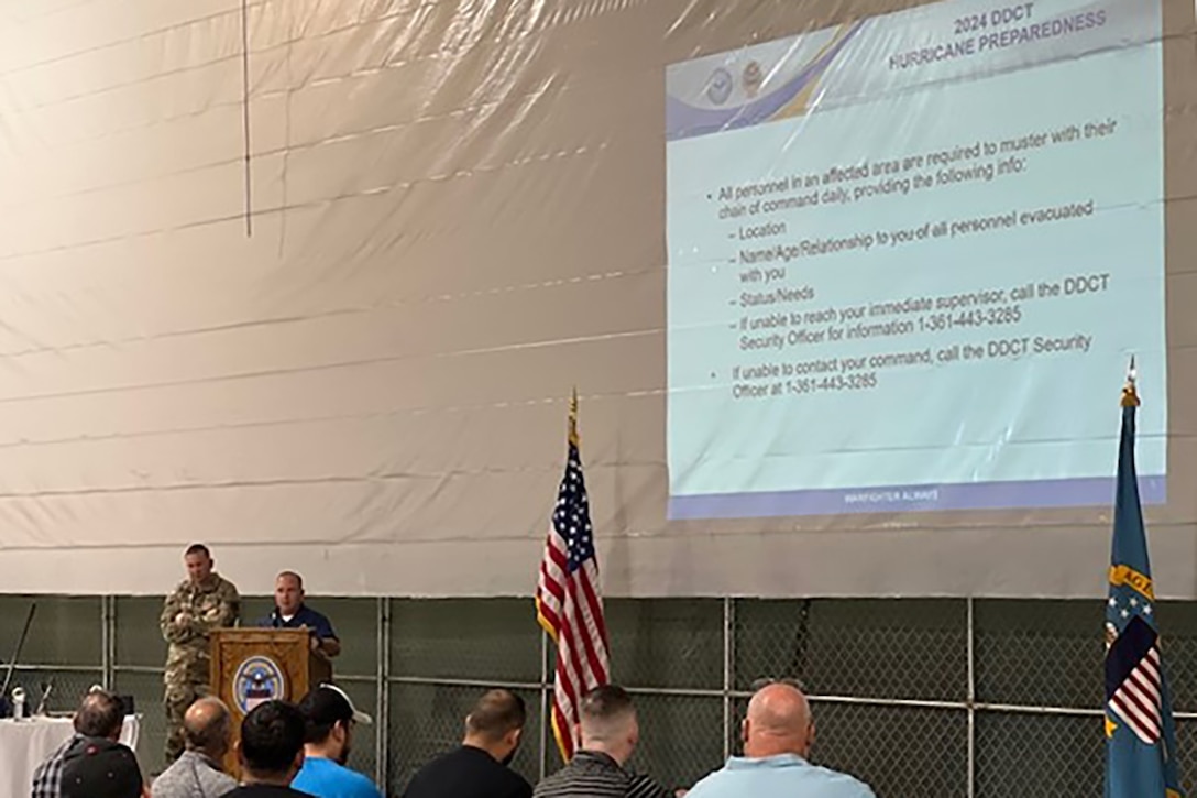 A man speaks to a group of seated people from behind a podium. There is a screen in front of the group that has hurricane safety information on it.