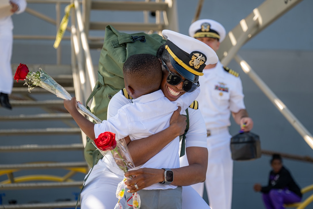 A sailor hugs a child during a homecoming event as both hold red roses.