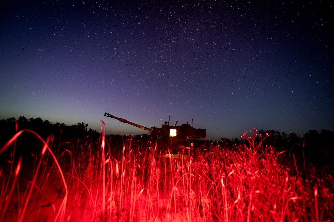 A military vehicle is parked in a grassy area at night, illuminated by red lights.