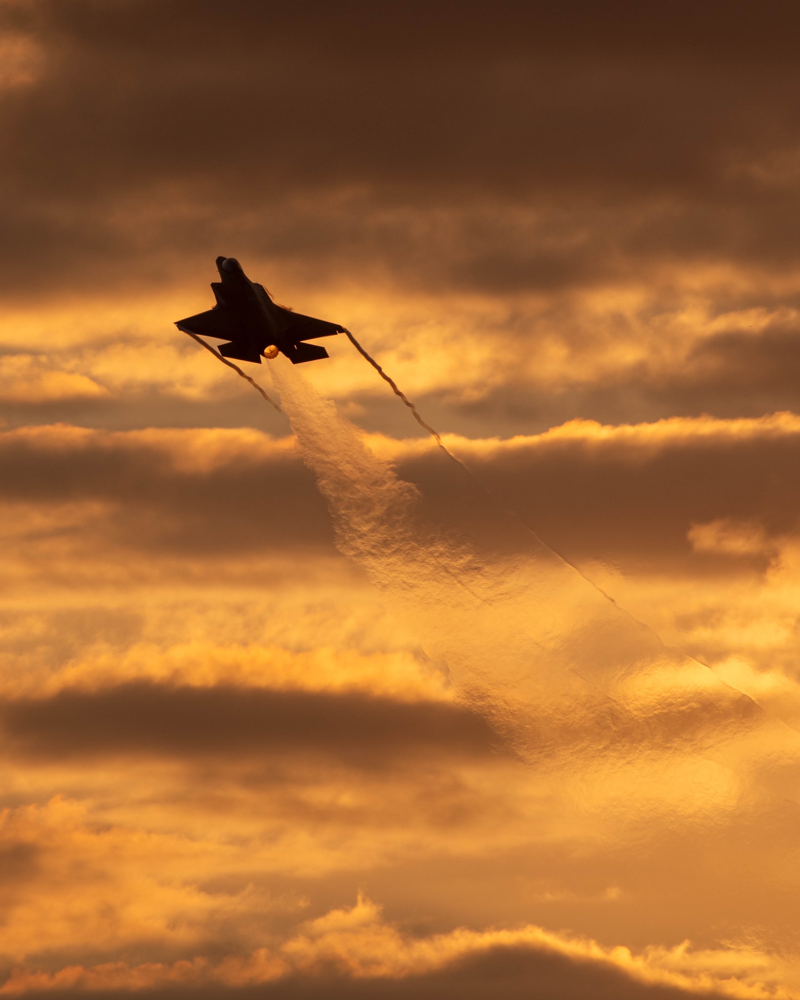 Silhouette of an F-35A flying into the sunset with vapor trail.
