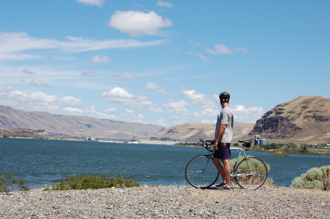 A man stands in front of his bicycle wearing a helmet in front of the John Day Dam.