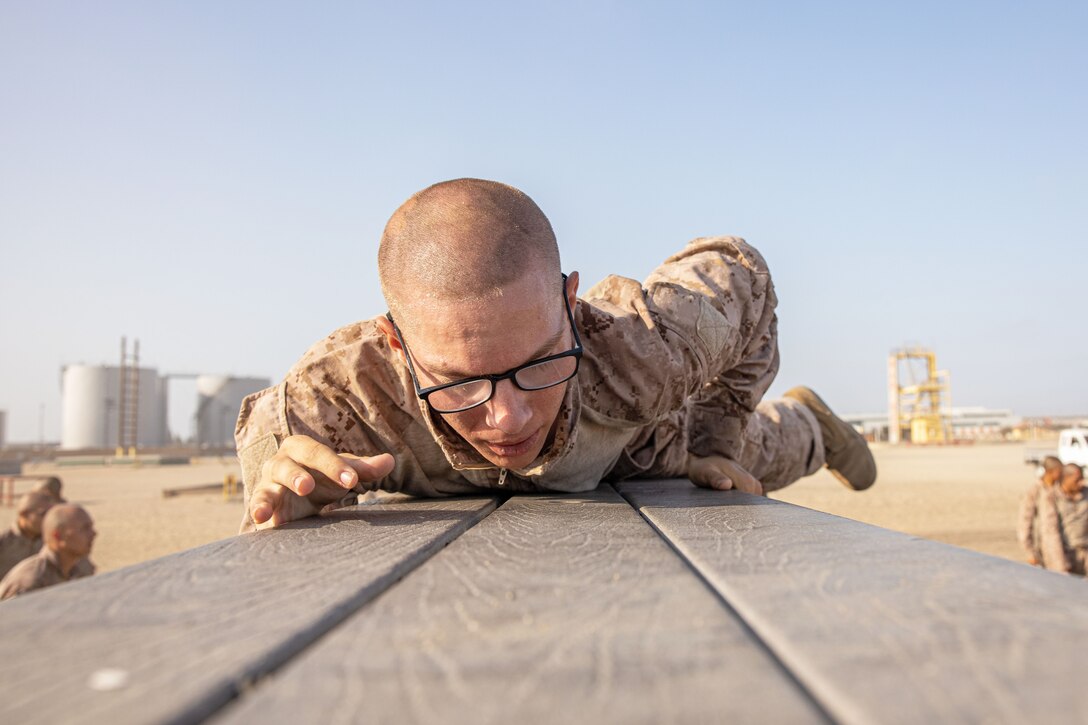 A close-up of a Marine Corps recruit climbing over an obstacle as fellow recruits watch in a desert-like area.