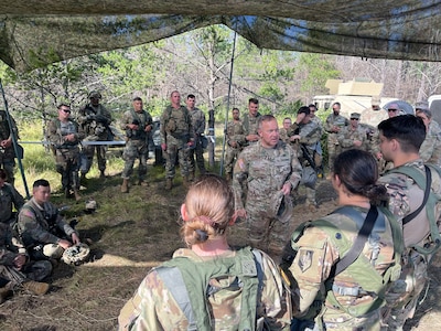 A group of uniformed soldiers stand in a semi-circle while listening to Lieutenant General Hale address them. He is standing in the middle speaking.