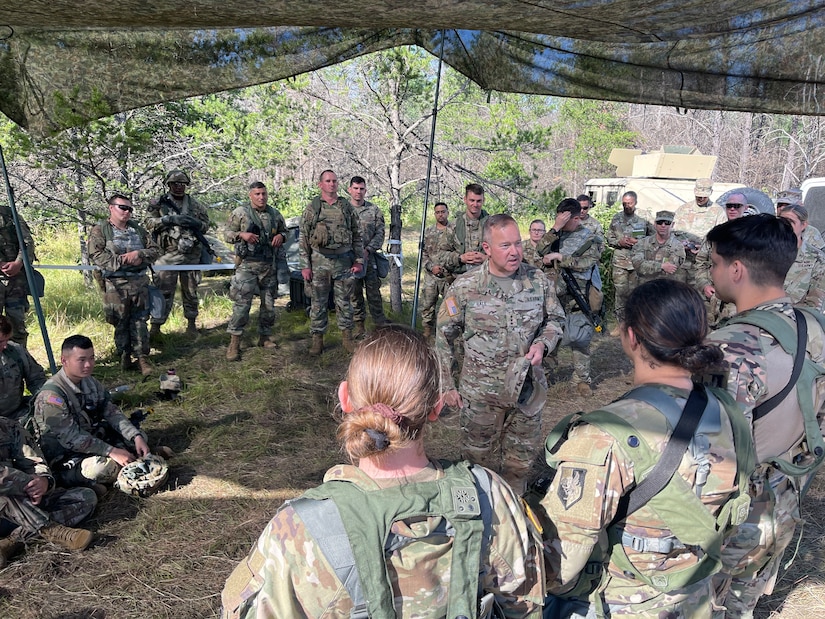 A group of uniformed soldiers stand in a semi-circle while listening to Lieutenant General Hale address them. He is standing in the middle speaking.