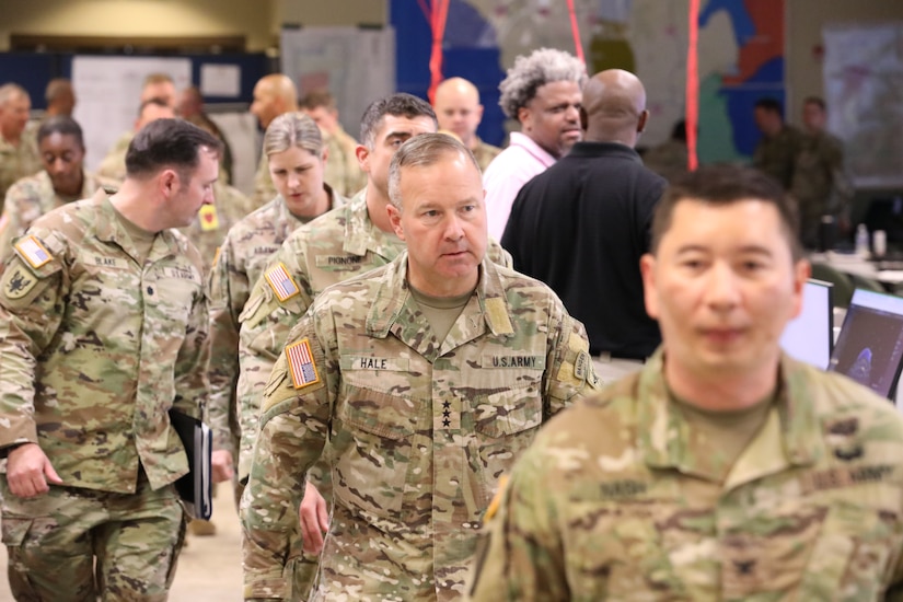 A column of soldiers in military uniform walk through an operations center.